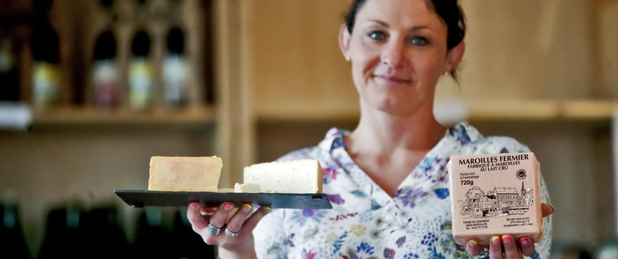 Joli jeune femme aux yeux bleus souriante robe fleuri, présentant dans sa main droite un plateau de fromage maroille, et dans sa main gauche un fromage maroille encore emballé